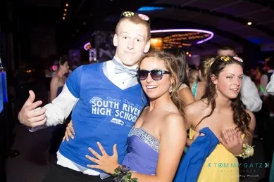 Young people in formal attire at a dance event, one wearing a "South River High School" shirt over a suit, with sunglasses and others in dresses.