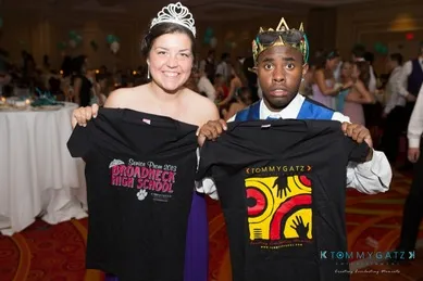 Two people wearing crowns hold up T-shirts at a formal event. The left shirt reads "Broadneck High School Prom Prince 2013," and the right shirt features yellow and red designs.