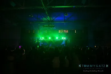 A dark venue with a crowd silhouetted against green stage lights, under a high ceiling with visible metal beams.
