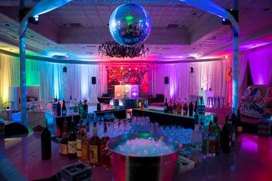 A vibrant, dimly lit party room with a disco ball, colorful lights, a chandelier, and a fully stocked bar with bottles and glasses.