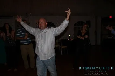 A man in a striped shirt stands with arms raised at a dimly lit event. People in the background watch him.