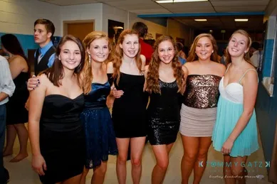 A group of six young women in dresses, smiling and posing together in a hallway at an indoor event.