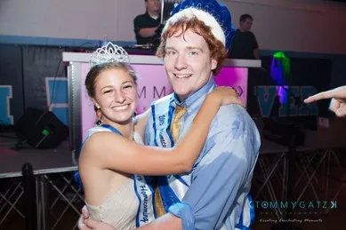 A young couple wearing crowns and sashes poses happily in front of a DJ booth at an event.