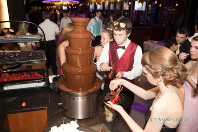 People enjoying a chocolate fountain at an event, dipping treats like strawberries and marshmallows. A server in a red vest and bow tie assists guests.