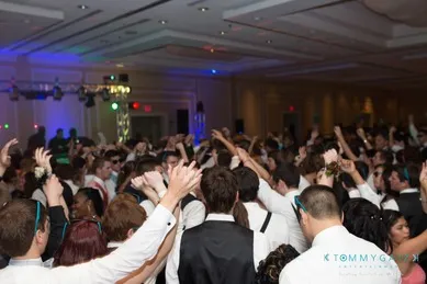 A crowded dance floor with people dressed in formal attire, raising their hands and dancing under colorful lights at an indoor event.