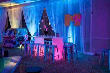A room with neon lights, white curtains, high tables with stools, and an MTV logo on the wall. A colorful painted van is parked nearby.