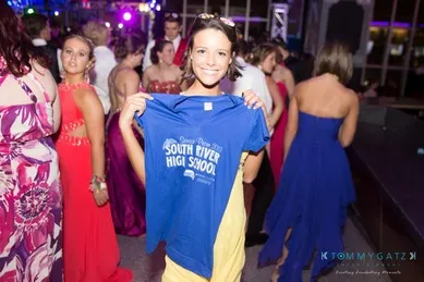 A person holds a blue "South River High School" T-shirt while standing at a social event. People in formal attire are mingling in the background.