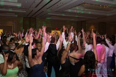 A large group of people in formal attire dancing with their hands in the air at an indoor event.