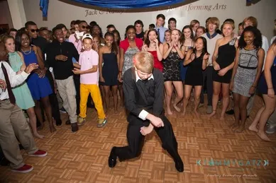 A person in formal attire dances energetically in the center of a room, surrounded by a group of people clapping and watching with smiles.