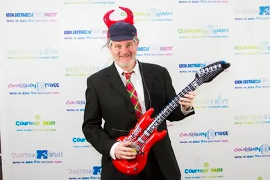 A man in a suit with a red horned hat holds an inflatable red guitar in front of a backdrop with repeated text and logos.