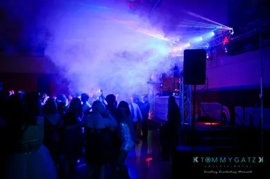A crowd dances at a dimly lit party with colorful lights and fog effects filling the room.