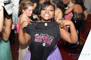 A young woman in a prom dress holds up a T-shirt reading "Senior Prom 2019 Broadneck High School." People are in the background at a dance event.