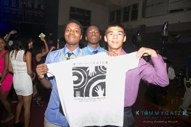Three young men in formal attire hold up a T-shirt at an indoor event, with people and party balloons visible in the background.