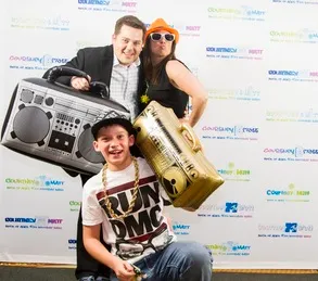 Three people posing with inflatable props in front of a branded backdrop. The man holds a gray boombox, the woman wears sunglasses and an orange hat, and the boy holds a gold radio.