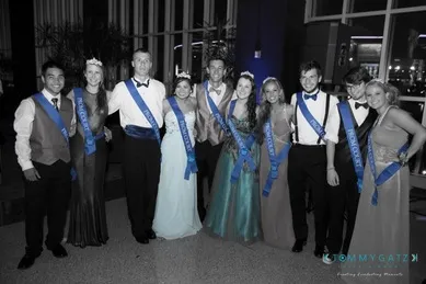 A group of ten people dressed formally, wearing blue sashes, posing indoors at an event. The background is dimly lit.
