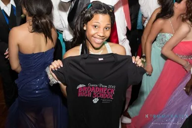 A person at a dance holds up a black T-shirt reading "Broadneck High School Senior Prom 2013," surrounded by others in formal attire.