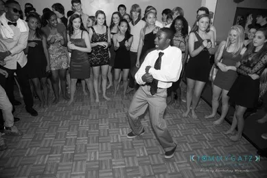 A person in a tie energetically dances in the center of a room while a crowd of people watch and react.