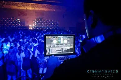 A DJ is performing at a crowded event with blue stage lighting, using a laptop to mix music.