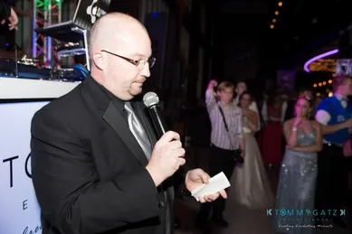 A man in a suit speaks into a microphone at an event, holding a folded piece of paper. People in formal attire stand in the background.