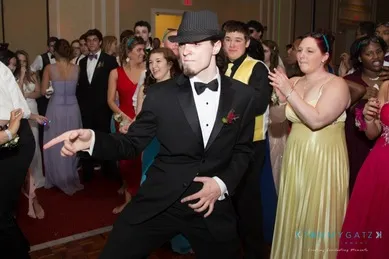 A person in a suit and hat dances energetically at a formal event, surrounded by people in gowns and tuxedos.