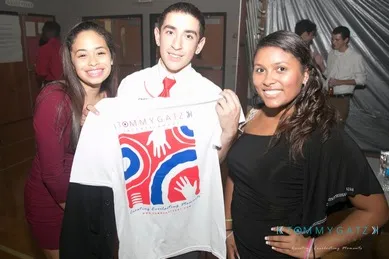 Three people smiling; the person in the center holds a brightly colored shirt with a design featuring hands.