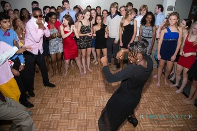 A group of people in formal attire stand around a person dancing energetically on a patterned floor.