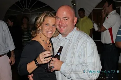 A smiling couple poses together at a party, holding drinks. Other people are in the background.