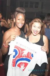 Two people smiling, holding a white t-shirt with a colorful design featuring stylized hands and shapes, in a crowded indoor setting.