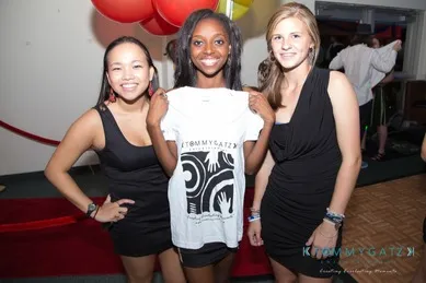 Three people stand together indoors. The person in the middle holds up a white T-shirt. The background features colorful balloons and a red carpet.