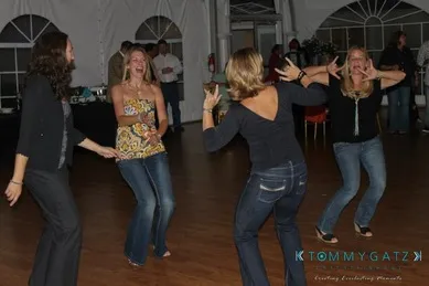 Four women dancing energetically in a dimly lit room with a wooden floor, laughing and enjoying the moment.