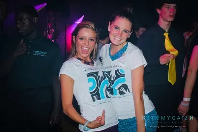 Two people smiling and posing at a party, wearing matching white t-shirts with blue patterns. Other people are in the background, including a person in a black shirt and another in a yellow tie.