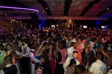 A large group of people in formal attire dance at a dimly lit event with colorful lighting.