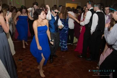 People in formal attire dancing energetically at a social event. A barefoot woman in a blue dress is prominent in the foreground.