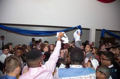 A large group of people are gathered indoors at an event, some raising their hands and holding napkins. Blue and red decorations hang from the ceiling.