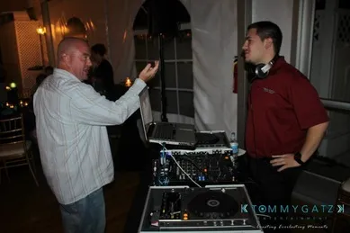 A man gestures while speaking to a DJ, who is standing behind a mixing console with headphones around his neck.