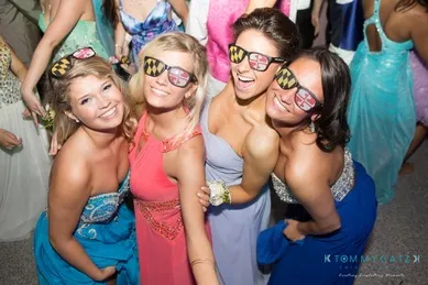 Four smiling women in formal dresses wearing novelty sunglasses with a distinctive red, black, and yellow pattern, posing closely together at an event.