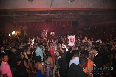 A large crowd of people dancing energetically in a dimly lit venue, with colored lights and raised hands visible.
