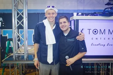 Two people standing side by side indoors, one wearing a white scarf and sunglasses, the other holding a microphone. They are both smiling.