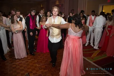 A group of people dressed in formal attire are dancing in a banquet hall. A person in a gold vest dances with a person in a pink dress in the center.