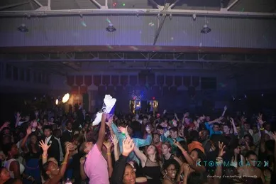 A large crowd of people dancing under dim lighting in a gymnasium, with raised hands and colorful clothing visible.