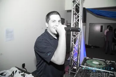 A man holding a microphone stands next to DJ equipment, preparing to speak or perform.