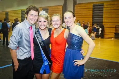 Four people smiling and posing together in a gym. One person wears a sash and crown.