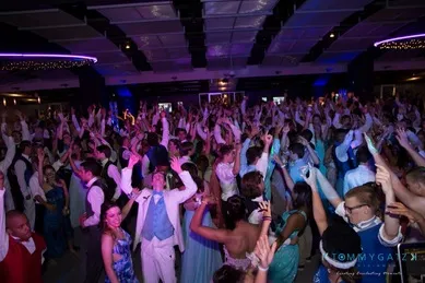 A large group of people in formal attire dance energetically at a crowded indoor event, with colorful lighting and hands raised in the air.