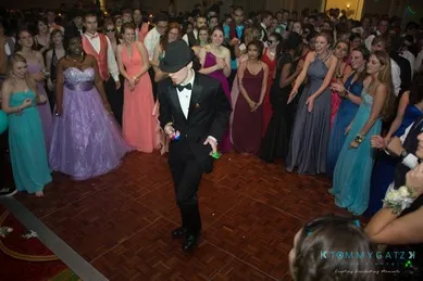 A person in a suit and hat dances on a crowded dance floor, surrounded by people in formal dresses and suits.