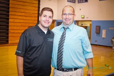 Two men standing together in a gymnasium, one wearing a black polo shirt and the other wearing a light blue dress shirt with a checkered tie.