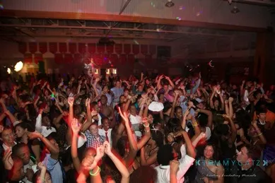 A large, energetic crowd is dancing and raising their hands in a dimly lit venue with colorful lights.