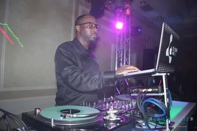A person wearing glasses is operating DJ equipment, including a laptop and turntables, in a dimly lit room with colorful lights.
