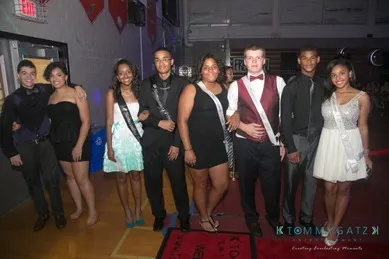 A group of eight people dressed in formal attire, standing in a gymnasium with banners hanging above.