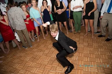 A person in a suit dances energetically on a wood-patterned floor, surrounded by people in formal attire.