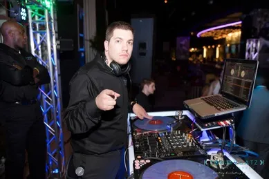 DJ standing at a turntable setup with a laptop, wearing headphones around the neck, points towards the camera. Security guard visible in the background.